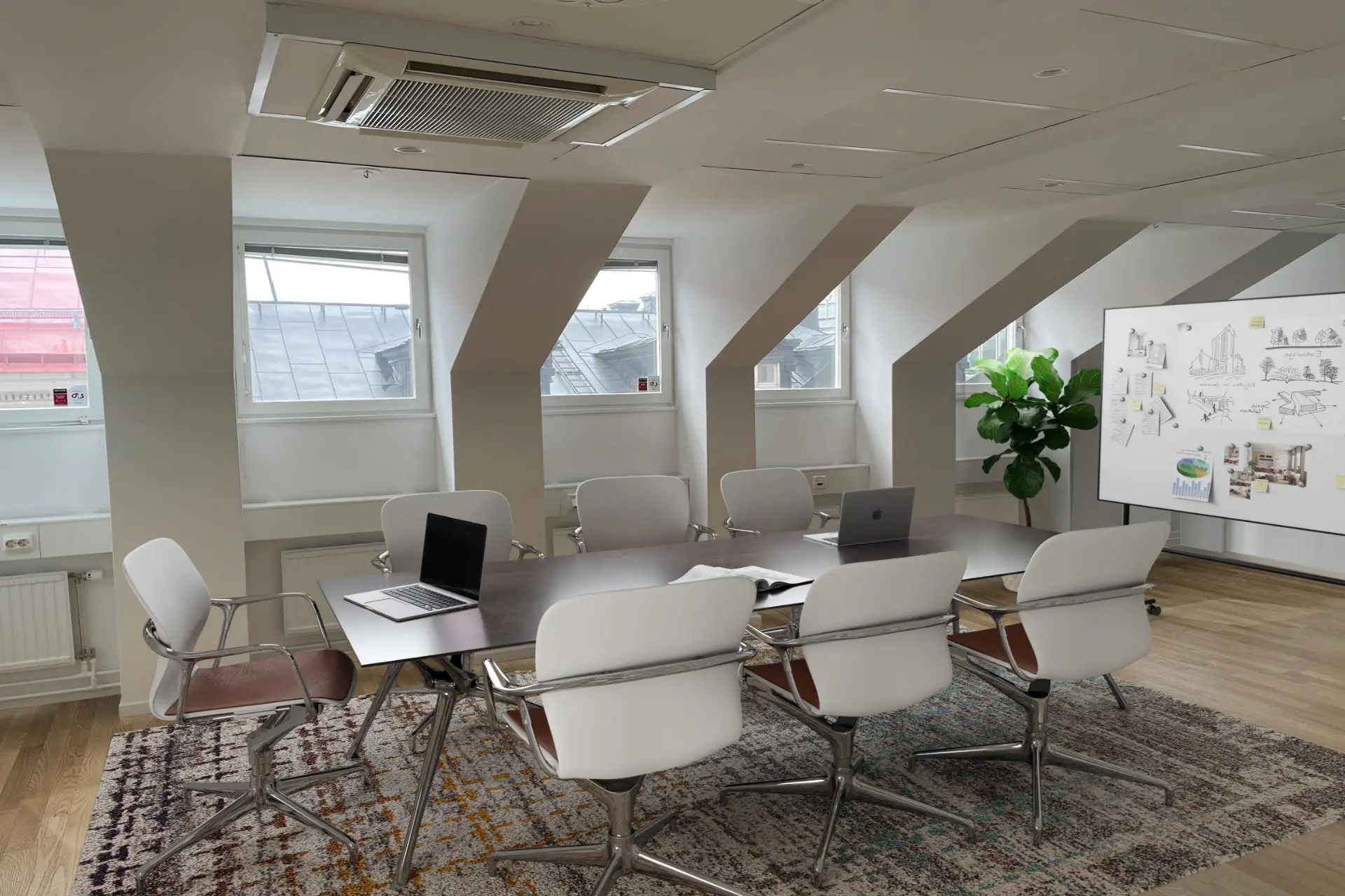 Kontorsmötesrum med sluttande tak, med ett stort mörkt bord, vita stolar, bärbara datorer och dekorativa växter, med gott om naturligt ljus från takfönster.